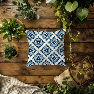 May include: A square throw cushion featuring a repeating diamond pattern in shades of blue and white. The design consists of intricate floral-inspired motifs centred within each diamond shape, set against a clean white background.