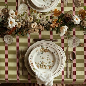 May include: A table setting on a green and white chequered tablecloth featuring white ceramic plates with floral patterns, silver forks, a silver knife, a silver spoon, clear glass tumblers, and a centrepiece of dried flowers and white blossoms.