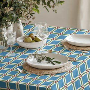 Puede incluir: Un mantel azul con un patrón geométrico de líneas blancas, verdes y rojas. La mesa está puesta con platos blancos, copas de vino y un cuenco de fruta. Los manteles individuales tejidos y la cubertería completan la escena, con un jarrón de flores al fondo.