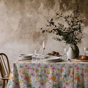 May include: A rectangular tablecloth featuring a vibrant, repeating pattern of tropical fruits, palm trees, and pink flowers on a light blue and white checkered background. The table is set with white ceramic plates, glassware, and a candle.