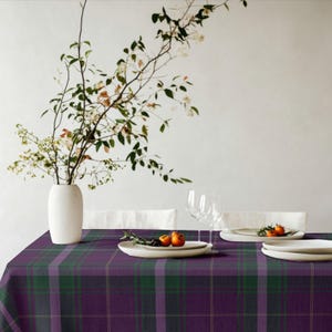 May include: A rectangular tablecloth featuring a purple and green plaid pattern with thin yellow grid lines. The fabric covers a table surface, with a white vase containing leafy branches and two white plates holding small orange fruits.