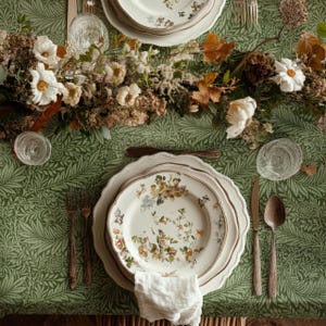 May include: A dining table set with floral plates, cutlery, and clear glasses. The green tablecloth features a leaf pattern. A floral arrangement of white flowers and autumn leaves adorns the table setting, creating an elegant atmosphere.