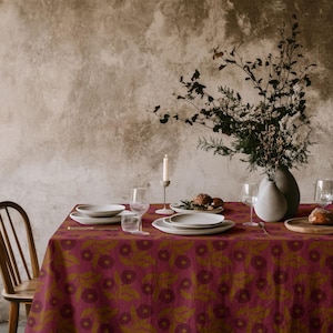 Puede incluir: Una mesa de comedor cubierta con un mantel magenta con un patrón floral oscuro repetitivo. La mesa está dispuesta con platos de cerámica blanca, copas de cristal transparente, una vela pequeña en un soporte gris y un jarrón con ramas secas.