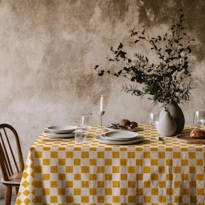May include: A mustard yellow and white checkered tablecloth covering a dining table. The table is set with white ceramic plates, clear glass stemware, a single white taper candle in a holder, and a vase containing dried branches and foliage.