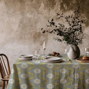 Op de afbeelding: Een eettafel gedekt voor een maaltijd, met een blauw tafelkleed met een bloemmotief in wit en groen. De tafel is gedekt met witte borden, glazen en een houten stoel. Een vaas met bloemen en een brandende kaars zijn ook aanwezig.
