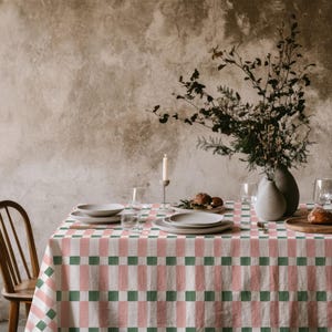 May include: A dining table set for a meal, featuring a tablecloth with a repeating pattern of pink and green squares. The table is set with plates, glasses, and a vase of flowers. A wooden chair is visible on the left.