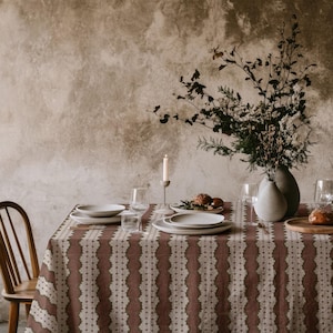 May include: A rectangular tablecloth featuring a deep brown base colour with vertical stripes of cream-coloured lace-like patterns. The table is set with white ceramic plates, clear glass stemware, a tall white candle, and a vase with dried branches.