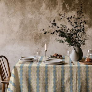 May include: A rectangular tablecloth featuring a vertical striped pattern in light blue and yellow on a cream background. The fabric is draped over a dining table set with white ceramic plates, clear glassware, a single candle, and a vase.