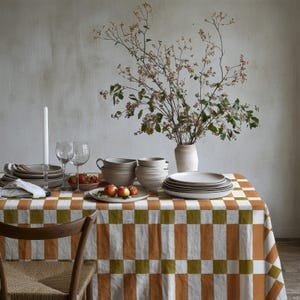 May include: A rectangular tablecloth featuring a repeating checkered pattern in alternating blocks of burnt orange, olive green, and white. The fabric is draped over a table set with ceramic bowls, plates, wine glasses, and a vase with branches.