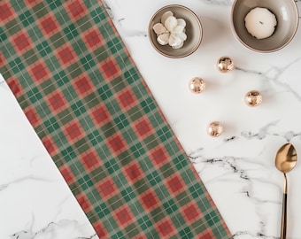 Pode incluir: Um caminho de mesa retangular longo com um padrão xadrez vermelho e verde. O caminho de mesa está posicionado ao lado de uma pequena tigela com uma flor branca, uma tigela maior com uma vela branca, três pequenas esferas douradas e uma colher dourada.