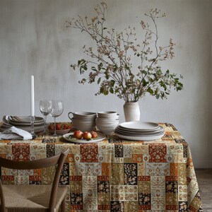 Puede incluir: Una mesa de comedor con un mantel estampado con cuadrados negros, marrones y dorados. La mesa está puesta con platos, cuencos, vasos y un jarrón con flores. También hay una vela encendida y manzanas.