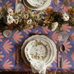 Op de afbeelding: Een tafelsetting met borden met bloemenpatroon, bestek en heldere glazen. Het blauwe tafelkleed heeft een roze bloemenpatroon. Een bloemstuk van witte en bruine bloemen en bladeren siert de tafel.