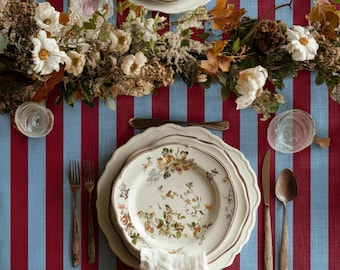 May include: A table setting featuring a cream-colored ceramic plate with a floral pattern, a white linen napkin, a silver fork, a silver knife, and a silver spoon, all arranged on a blue and red striped tablecloth with a dried floral centerpiece.