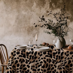 Könnte beinhalten: Eine rechteckige Tischdecke mit einem auffälligen Leopardenmuster in Schwarz und Beige. Der Stoff bedeckt einen Esstisch, der mit weißen Keramiktellern, klaren Weingläsern, einer weißen Kerze im Halter und einer Vase mit Grünpflanzen gedeckt ist.