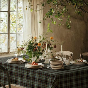 May include: A dining table covered with a dark green and blue plaid tablecloth, featuring a large floral centerpiece with orange and yellow flowers, white taper candles in holders, ceramic plates, bowls, and clear glassware arranged for a meal.