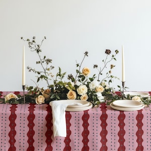 May include: A table setting featuring a pink and dark red patterned tablecloth, a central floral arrangement of cream and peach roses with green foliage, two tall cream taper candles in dark holders, and stacked white ceramic plates with a napkin.