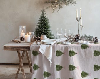 May include: A light-colored tablecloth featuring a repeating pattern of green Christmas trees with small pink bows. The fabric drapes over a wooden table set with white plates, clear wine glasses, white candles in glass holders, and pinecones.