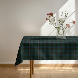 May include: A rectangular tablecloth featuring a dark green, navy blue, and thin orange plaid pattern. Two clear glass vases sit on the surface, containing a bouquet of pink carnations and small white flowers with green stems and leaves.