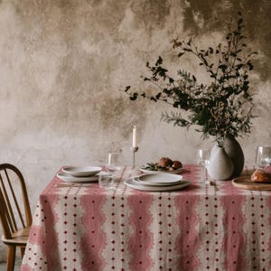 May include: A rectangular tablecloth featuring a repeating pattern of vertical dusty rose bands with scalloped edges and small dark red star motifs on a cream background, draped over a table set with white ceramic plates and glassware.