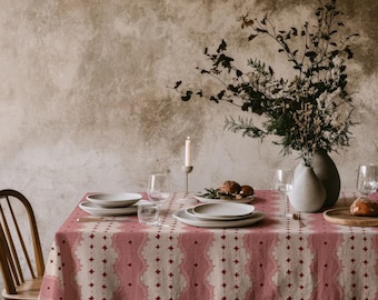 May include: A rectangular tablecloth featuring a repeating pattern of vertical dusty rose bands with scalloped edges and small dark red star motifs on a cream background, draped over a table set with white ceramic plates and glassware.