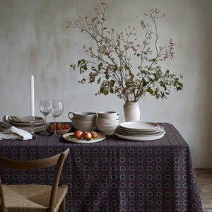 Puede incluir: Una mesa de comedor decorada con un mantel estampado en burdeos, verde y blanco. La mesa está puesta con platos, cuencos, vasos y un jarrón con flores. Una silla de madera es parcialmente visible.