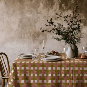 Puede incluir: Una mesa de comedor preparada para una comida, con un mantel con un patrón repetido de cruces verdes, blancas y rosas. La mesa está puesta con platos, vasos y un jarrón con flores. También se ven una vela encendida y una silla de madera.