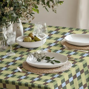 May include: A table setting featuring a tablecloth with a green, brown, and light blue checkered pattern. The table includes white ceramic plates, clear glass wine glasses, a white bowl filled with green pears, and a vase with greenery.