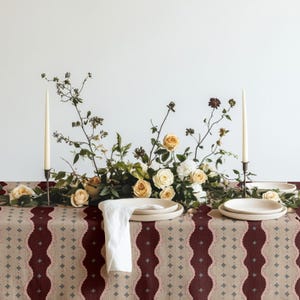 Könnte beinhalten: Ein gedeckter Tisch mit einer Tischdecke, die ein vertikales Muster aus dunkelroten und beigen Wellenstreifen mit kleinen grauen Rauten zeigt. Ein Blumenarrangement aus cremefarbenen und pfirsichfarbenen Rosen steht zwischen zwei dünnen Kerzen.