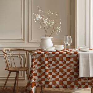 May include: A rectangular tablecloth featuring a repeating geometric pattern of burnt orange and white semicircles. The fabric drapes over a table set with a white pitcher containing white flowers, two clear glass flutes, and white bowls.
