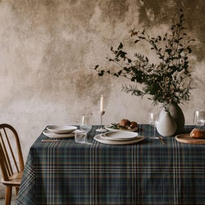 May include: A dining table covered with a dark green, navy blue, and thin orange plaid tablecloth. The table is set with white ceramic plates, clear glass stemware, a tall white candle in a holder, and a vase containing dried branches.