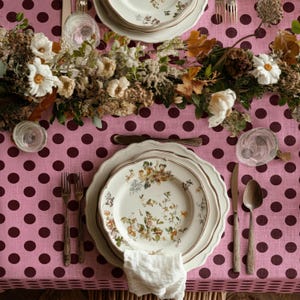 May include: A table setting with floral patterned plates, silverware, and a wooden spoon. The table is covered with a pink polka dot tablecloth and decorated with a floral centerpiece. Clear glasses are placed around the plates.