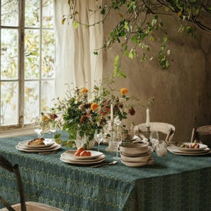 May include: A dining table covered with a dark teal patterned tablecloth, set with white ceramic plates, bowls, and glassware. A large floral centerpiece with orange and yellow flowers sits in the middle, accompanied by two white taper candles.