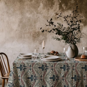 May include: A rectangular tablecloth featuring a repeating pattern of stylised blue and muted red floral motifs on a cream background. The table is set with white ceramic plates, clear glassware, a tall candle in a holder, and a vase with dried branches.