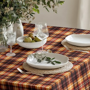 May include: A rectangular tablecloth featuring a plaid pattern in shades of orange, brown, black, and white. The table is set with white ceramic plates, woven placemats, clear glass stemware, and a white bowl containing green pears.