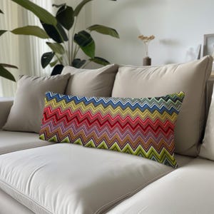 Puede incluir: Un cojín lumbar rectangular con un vibrante patrón de chevrón multicolor. El diseño consiste en rayas horizontales en zigzag en tonos azul, amarillo, rojo, rosa y verde, creando un motivo geométrico repetitivo en toda la superficie.