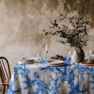 May include: A white tablecloth featuring a repeating blue scrollwork pattern draped over a dining table. The table is set with white ceramic plates, clear glass stemware, a single lit candle in a holder, and a vase filled with dried branches.