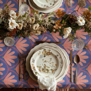 May include: A table setting featuring a blue tablecloth with a pink fan pattern, white floral plates, a white napkin, two forks, a knife, a spoon, and two clear glasses. A centrepiece of dried flowers and white blossoms runs across the table.