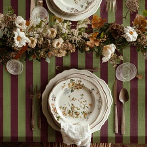May include: A table setting featuring a white ceramic plate with a floral pattern, placed on a dark red and green striped tablecloth. The setting includes a silver fork, a silver knife, a silver spoon, a clear glass, and a white cloth napkin.