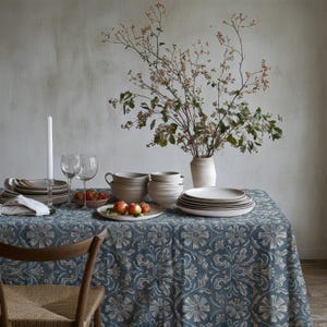 May include: A blue and white floral patterned tablecloth covers a table set with a stack of ceramic plates, nested bowls, two wine glasses, a white taper candle in a holder, a small bowl of red berries, and a vase with leafy branches.