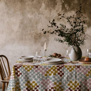 Puede incluir: Un mantel rectangular con un patrón repetitivo de pequeños cuadrados de estilo pintado a mano en tonos apagados de verde oliva, rosa empolvado, azul claro y granate oscuro, que cubre una mesa de comedor con platos y cristalería blanca.