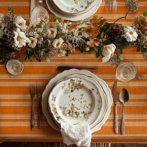 May include: An elegantly set table featuring floral patterned plates, silverware, and a white linen napkin. The orange striped tablecloth is decorated with a floral centerpiece and glassware, creating a refined dining setting.