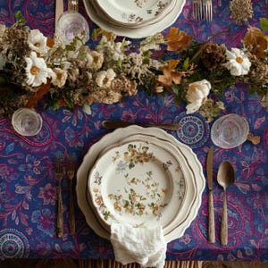 May include: A table setting featuring floral plates, cutlery, and a white napkin. The blue paisley tablecloth is adorned with a floral centrepiece. The plates have a gold trim. The scene evokes a sense of elegance.