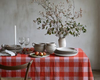 May include: A red and light pink checkered tablecloth covers a table set with a stack of white ceramic plates, nested bowls, two clear glass wine glasses, a tall white candle, and a small plate of apples next to a vase with leafy branches.