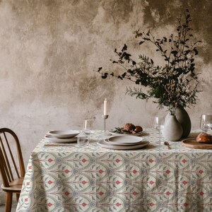 May include: A rectangular tablecloth featuring a repeating pattern of stylised floral motifs in shades of red, blue, and green on a cream background. The fabric drapes over a table set with white ceramic plates, glassware, and a vase of dried branches.