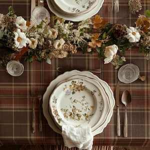May include: A table setting featuring a brown and tan plaid tablecloth, white scalloped plates with floral patterns, clear glass tumblers, metal cutlery, and a centerpiece of dried white flowers, brown leaves, and greenery arranged on the table.