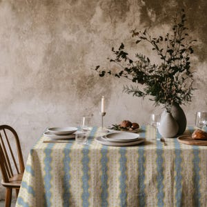 Könnte beinhalten: Ein Esstisch, der für eine Mahlzeit gedeckt ist, mit einer blau-gelb gemusterten Tischdecke. Der Tisch ist mit Tellern, Gläsern und einem hölzernen Brotbrett gedeckt. Eine Vase mit Blumen und eine brennende Kerze tragen zum Ambiente bei.