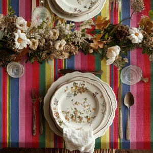 May include: A dining table set with floral plates, cutlery, and a white napkin. The table is covered with a colourful striped tablecloth. A floral arrangement with white flowers and greenery runs down the centre of the table.