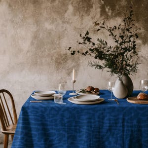 May include: A vibrant blue tablecloth with a subtle, wavy tonal pattern covers a dining table. The table is set with white ceramic plates, clear glass stemware, a single tall white candle in a holder, and a vase filled with dried branches.