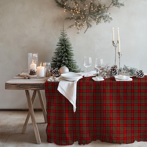 Könnte beinhalten: Ein festlich gedeckter Esstisch für eine Weihnachtsfeier. Der Tisch ist mit einer rot-grünen karierten Tischdecke bedeckt. Weiße Teller, Servietten und Weingläser sind arrangiert. Kerzen, ein kleiner Weihnachtsbaum und Tannenzapfen ergänzen die festliche Dekoration.