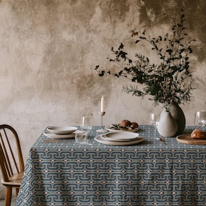 Puede incluir: Una mesa de comedor con un mantel estampado en azul y rosa. La mesa está puesta con platos blancos, vasos y un jarrón con flores. Una silla de madera es visible a la izquierda. La mesa está lista para una comida.